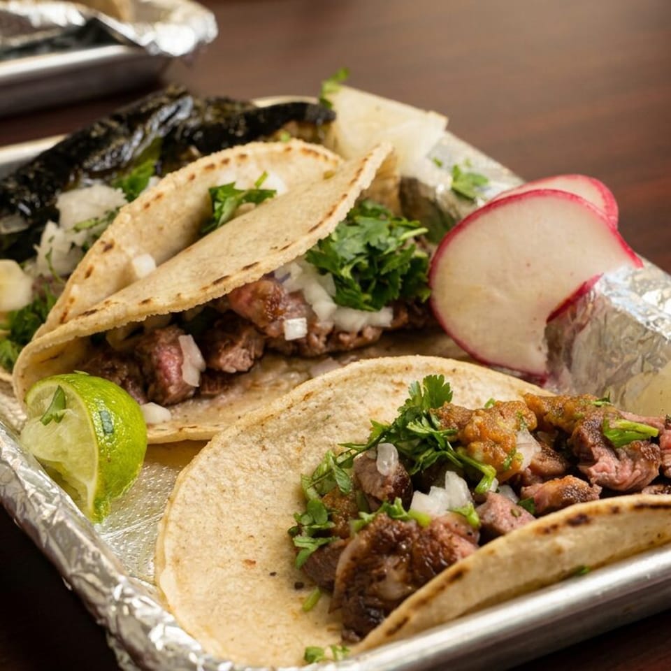 Four soft corn tacos filled with grilled meat, onions, cilantro, and salsa, served with lime and radish on a foil-lined tray.