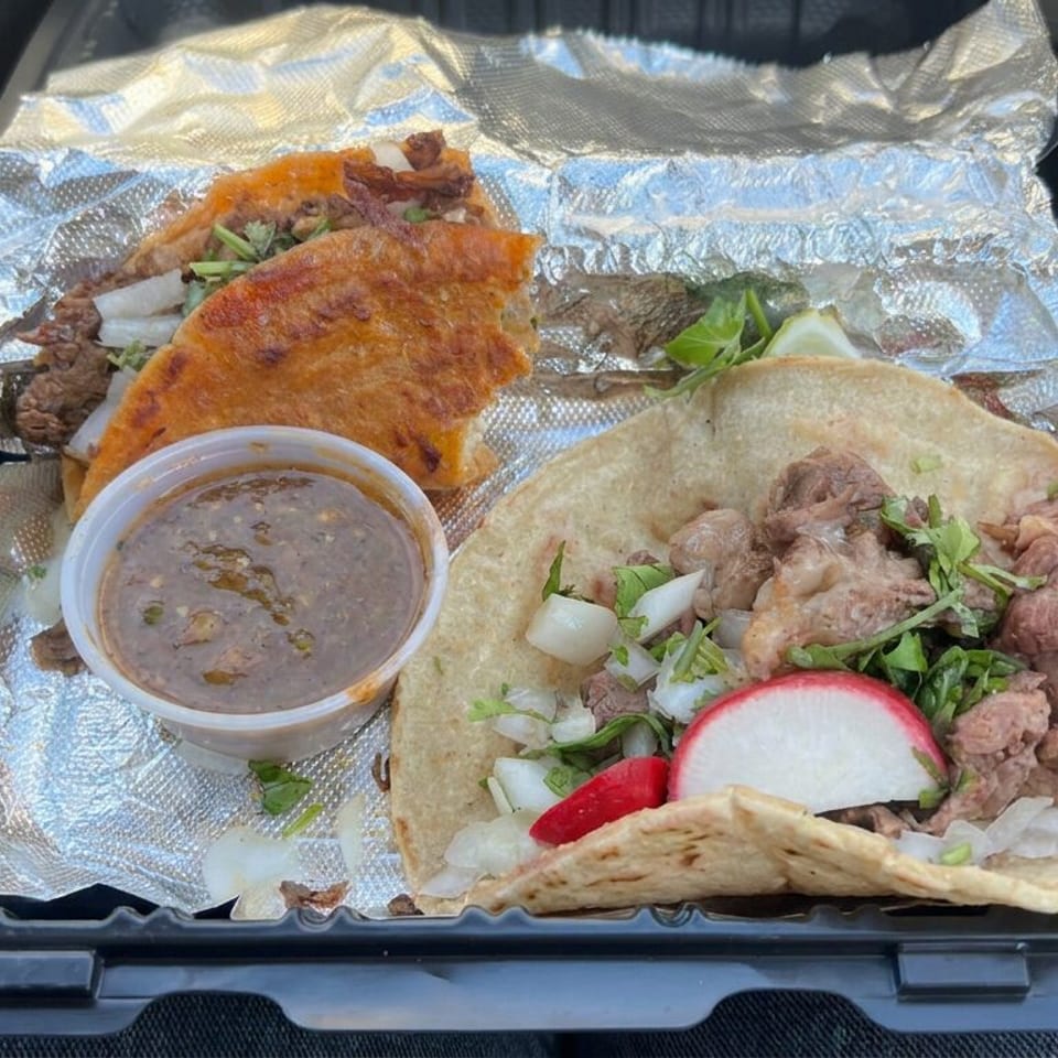Takeout tray holding meat tacos with onions, cilantro, radishes, a crispy taco, and a container of dipping sauce.
